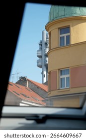 View through a window frame showing a mix of architectural styles: a rounded yellow building with a green dome, red-tiled roofs, and a modern telecommunications tower in the background. Clear blue sky