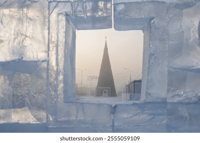 View through a window in an ice wall to a New Year tree. Winter view of a large Christmas tree on a snowy city square. Cold weather. In front of the tree there is a sign in Russian 