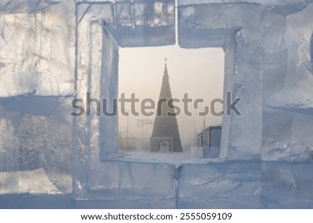 View through a window in an ice wall to a New Year tree. Winter view of a large Christmas tree on a snowy city square. Cold weather. In front of the tree there is a sign in Russian 