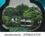 A view through a window of a Suzhou garden, showcasing a pond with lily pads and a traditional Chinese pavilion nestled amongst lush greenery.