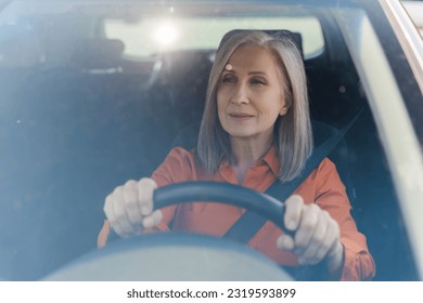 View through the windshield, portrait of a gray-haired caucasian senior woman 60s, female taxi driver, holding hands on the steering wheel, driving a car with a fastened seat belt. Road insurance. - Powered by Shutterstock