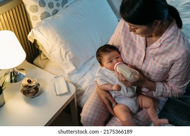 view from top asian mother wearing pajamas is nursing her adorable newborn baby with bottled milk by the bedside table in a cozy bedroom with lamp light. - Shutterstock ID 2175366189
