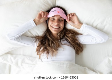 View from top happy attractive woman taking off pink sleep mask having charming toothy wide smile pose look at camera feels refreshment after enough night sleep, nap lying in home or hotel bed concept