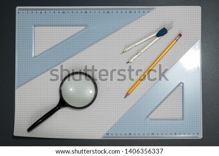 View from the top of scholastic material positioned on a background of squares, visible pencil, a magnifying glass, a compass, two drawing brackets. Back to school concept.