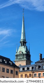 View of a cathedral’s tower and spire, a striking example of Gothic architecture with a copper-green roof and ornate detailing. The clock face with Roman numerals is visible on the tower, and the spire rises sharply into a vivid blue sky, creating a dramatic vertical composition.