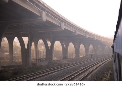 View from a train of tracks passing under a massive concrete overpass with arches in a hazy, misty morning.