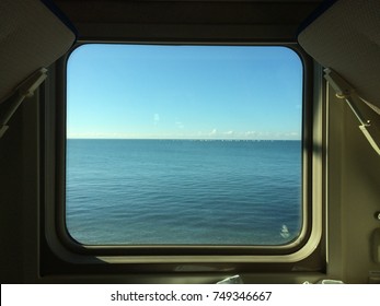 View from the train window to the calm blue sea. The sea merges with the blue clear sky on the horizon. A flock of gulls sits on the water in the distance. 