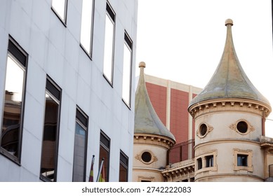 View of two distinct architectural styles in Barcelona. A historical building with two towers stands beside a modern structure. The scene shows the blend of old and new in city design.