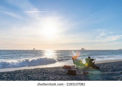 View Of Two People In Chairs On Rialto Beach On Coastal Stretch Of Olympic National Park, WA, USA With Lens Flare Of Late Afternoon Low Sun And Large Number Of Sea Stack Rock Formations In The Ocean