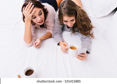 Up View Two Young Attractive Women Having Fun Together On White Bed. Good Morning Of Pretty Girls, Surfing In Internet On Phone, Drinking Tea, Smiling