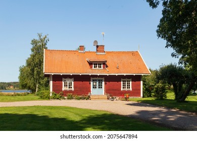 View Of A Typical Wooden Red Swedish House With White Window Frames On A Green Grassy Lawn On A Sunny Summer Day.