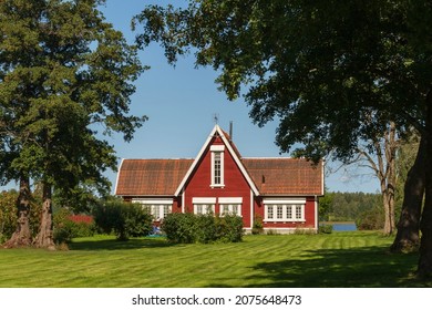 View Of A Typical Wooden Red Swedish House With White Window Frames On A Green Grassy Lawn On A Sunny Summer Day.