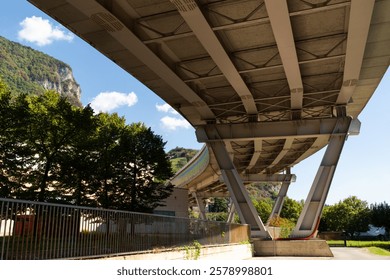 View from underneath large bridge with visible support beams. The scene blends urban infrastructure with the surrounding natural environment