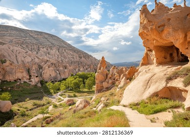 A view of unique cave formations and high rock hills in a valley. Green trees line the path leading through the landscape. The scene captures a sunny day with clouds.