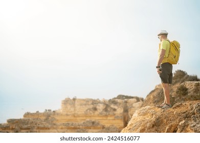 View of an unrecognizable man with backpack and hat standing ,watching the sunset from a cliff by the sea, concept of leisure and relax, copy space for text. High quality photo