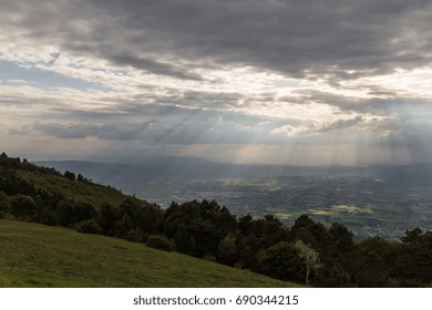 View of a valley from a mountain, with sun rays coming out through the clouds and lighting up some areas of the ground 