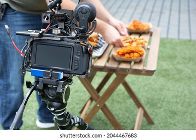 A View Of A Video Camera In Front Of A Patio Table Of American Street Food, In An Outside Patio Setting. A Person Styles The Entrees.