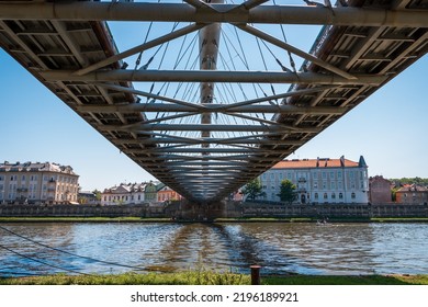 View Of The Vistula River From Under The Footbridge. Beautiful Historic Tenement Houses On The Other Side Of The River. The Steel Structure Of The Bridge. Cracow, Poland