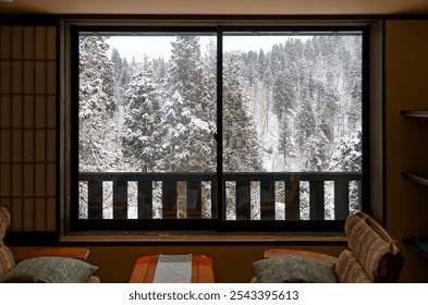 View from a warm cozy living room looking out thru the window toward the snowy winter landscape of snow covered forests, with chairs and a small table by the wooden deck, in a Japanese Onsen Ryokan - Powered by Shutterstock