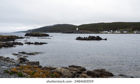 The view from Wester Head spans across the rugged bay of Hant's Harbour, leading to the white lighthouse on the Bay de Verde Peninsula in the Avalon region of Newfoundland, Canada, under a gloomy sky.
