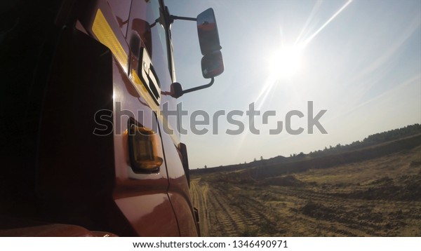 View from\
a wheel of the off-road truck riding in a dirt road on cloudy sky\
background. Scene. Close up for red cab of a lorry moving on the\
country, dusty road in a summer, sunny\
day.