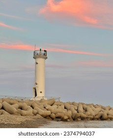 A view of the white lighthouse on the breakwater. The thin clouds behind the lighthouse are sunset, colored with pastel colors of light pink and orange medium tones.