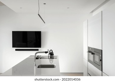 View of white modern kitchen with black TV hanging on white empty wall, counter with faucet and sink, induction stove top with kettle on it, cupboard with built in oven and microwave - Powered by Shutterstock