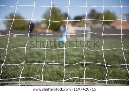 View of a white soccer football net with the blurred green field and people playing sport on a bright summer day