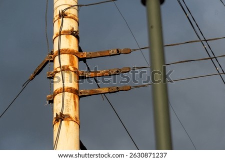 A view of a white utility pole exhibiting significant rust around its metal supports and weathered cables extending from it. The cloudy gray sky provides a somber background. 
