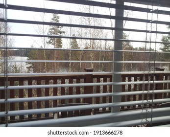 View from the window with blinds to the lake. Autumn day. White gray sky with clouds. Forest landscape. Window frame grill. There are horizontal blinds on the window. Outside the window there is a woo