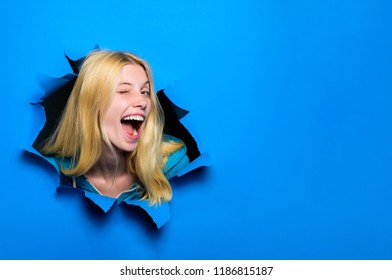 View of woman face through hole in blue paper. Winking smiling girl making hole in paper. Fashion and beauty concept. Copy space for advertising, to insert text or slogan. Discount, sale, season sales