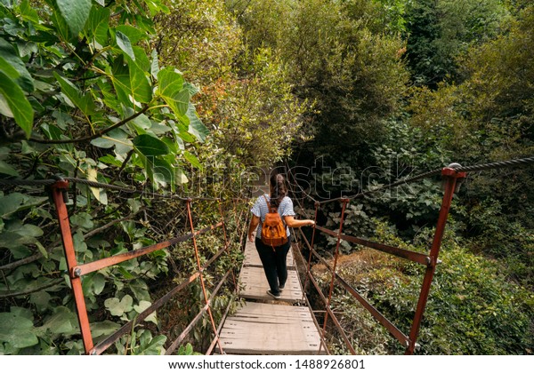view of a woman with a leather backpack\
crossing a hanging bridge into the\
forest.