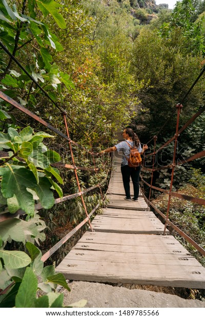 view of a woman with a leather backpack\
crossing a hanging bridge into the\
forest.