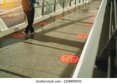 ฺBack view of woman walking on pedestrian glass bridge. Corridor connections between buildings with social distancing symbols on floor for distance footstep. Footsteps sticker stamp on bridge floor.