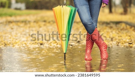A view of a woman's legs in rubber boots standing in a puddle, leaning on an umbrella. A woman standing in a puddle surrounded by fallen autumn leaves.