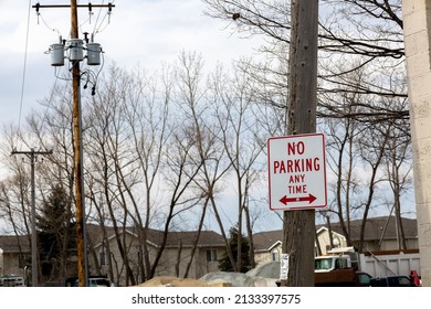 View Of Wooden Utility Pole And Housing With No Parking Sign Posted. Other Poles With Transformers Attached. Bare Tree Branches With Dump Truck And Snow Pile. Blue Sky With Fluffy Clouds. 