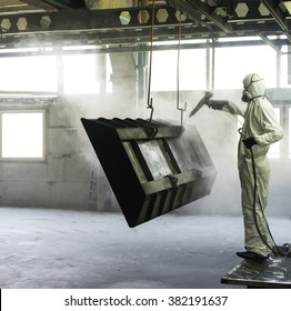 view of a worker wearing a full white protective suit and breathing mask, sand blasting a metal crate hung from a metal beam in the ceiling of an industrial hall - Powered by Shutterstock