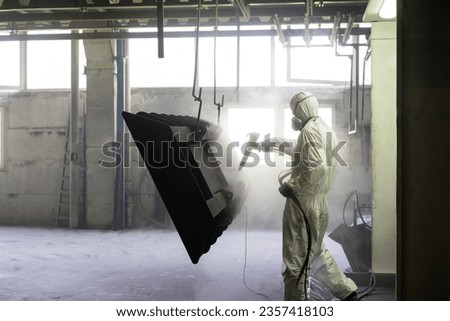 view of a worker wearing a full white protective suit and breathing mask, sand blasting a metal crate hung from a metal beam in the ceiling of an industrial hall