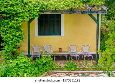 View Of Yellow House Wall And Terrace In The Garden – White Chairs, Table, Blooming Flowers, Green Bushes And Grass.