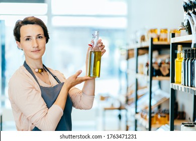 View of a young attractive woman working in a grocery store, female owner holding glass bottle with olive oil over shelves with eco packaging free products working in a zero-waste shop. - Powered by Shutterstock