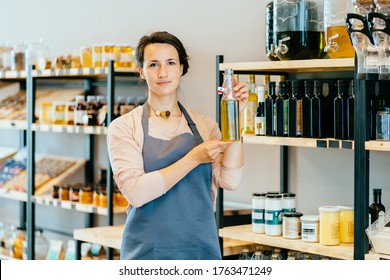 View Of A Young Attractive Woman Working In A Grocery Store, Female Owner Holding Glass Bottle With Olive Oil Over Shelves With Eco Packaging Free Products Working In A Zero-waste Shop.