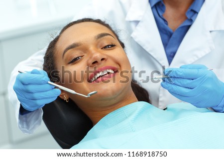 View of young beautiful afro woman with beautiful white teeth sitting in dental chair, smiling while dentist cures her teeth. Dentist wearing in latex gloves and holding instruments in hand.