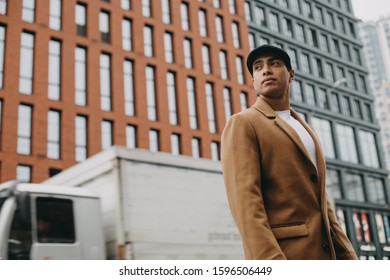 View Of Young Handsome Arabian Man Stand Outside And Pose. Truck Driving Behind Him. Guy Look Forward. Calm Peaceful And Cocentrated. Modern Urban Buildings Behind