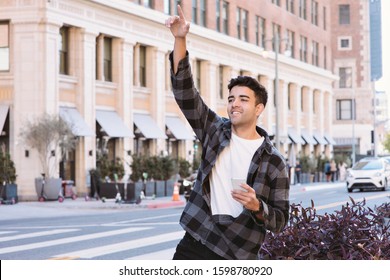 View Of A Young Latin Man Hailing A Ride Share Car With A Cell Phone In The City - Afternoon With Bright Lighting