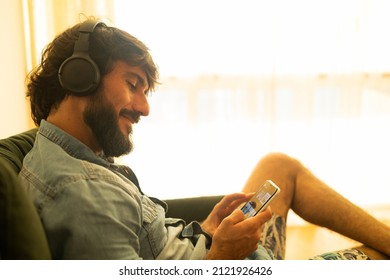 View Of Young Man Listening To Music With Headphones In His Ears In The Living Room Of His Apartment. High Quality Photo. Mobile Phone, Technology, Music, Urban Living, Feel Good Concept.