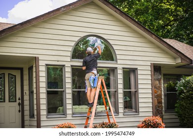 View Of Young Man Window Washer Stands On Ladder Holding Squeegee While Washing Outside Arch Window Of Midwestern House In Summer 