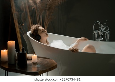 View of a young woman lying in a bath with foam and listening to music on headphones. Beautiful woman takes a relaxing bath to relieve stress after a hard day. - Powered by Shutterstock