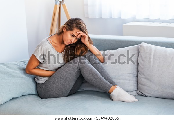 View of\
young woman suffering from stomachache on sofa at home. Woman\
sitting on bed and having stomach ache. Young woman suffering from\
abdominal pain while sitting on sofa at\
home