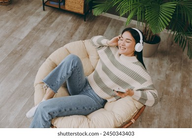 To view young woman wears casual clothes listen music in headphones use mobile cell phone sit on armchair stay at home flat rest relax spend free spare time in grey living room indoor. Lounge concept - Powered by Shutterstock