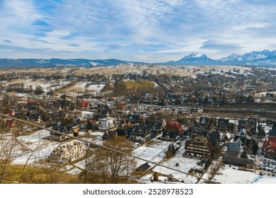 A view of Zakopane, Poland, reveals alpine-style buildings spread across a valley. Snow-covered Tatra peaks rise in the background, set against a cloudy sky - Powered by Shutterstock